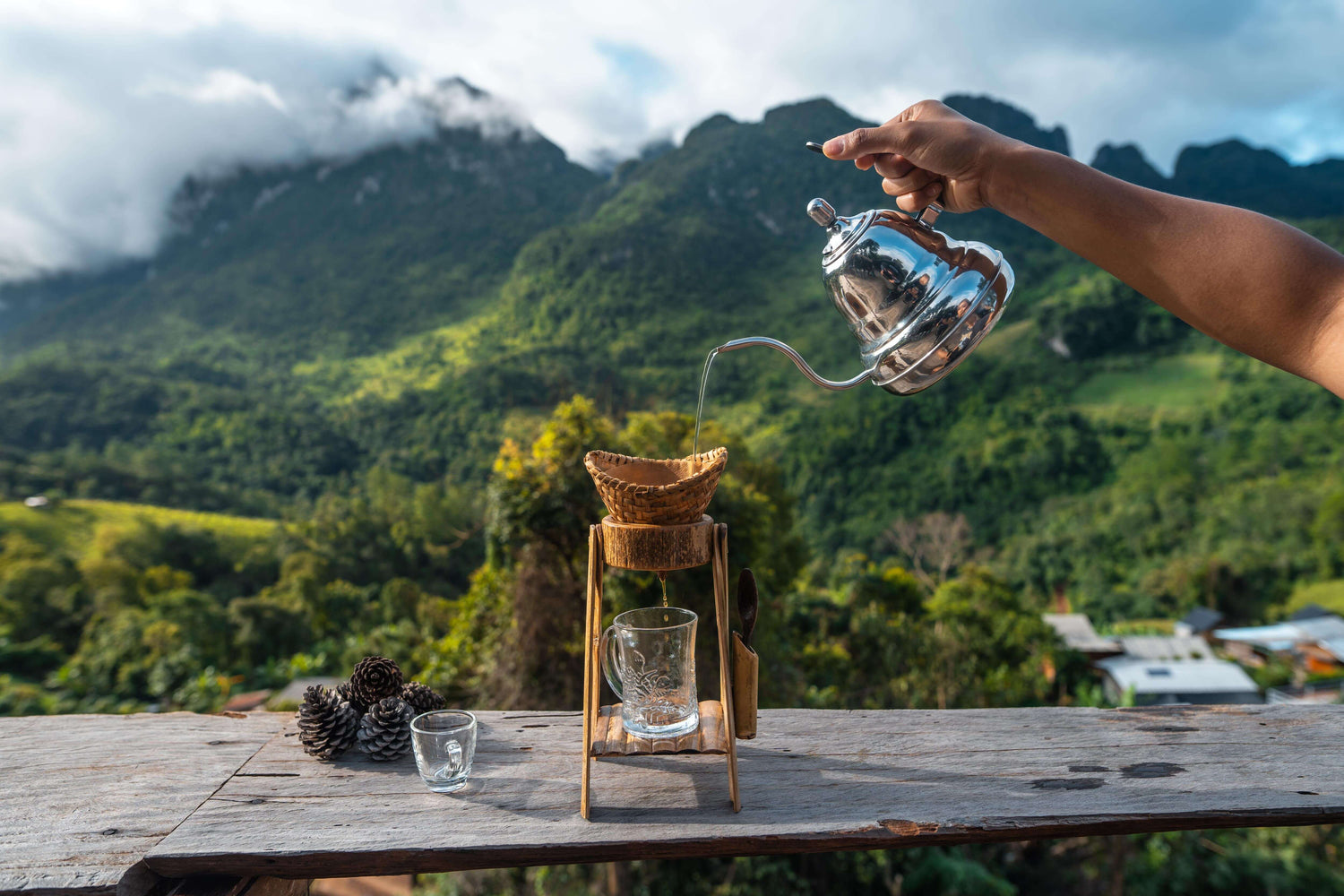 Hand pouring hot water from gooseneck kettle into pour-over coffee dripper with scenic mountain landscape background