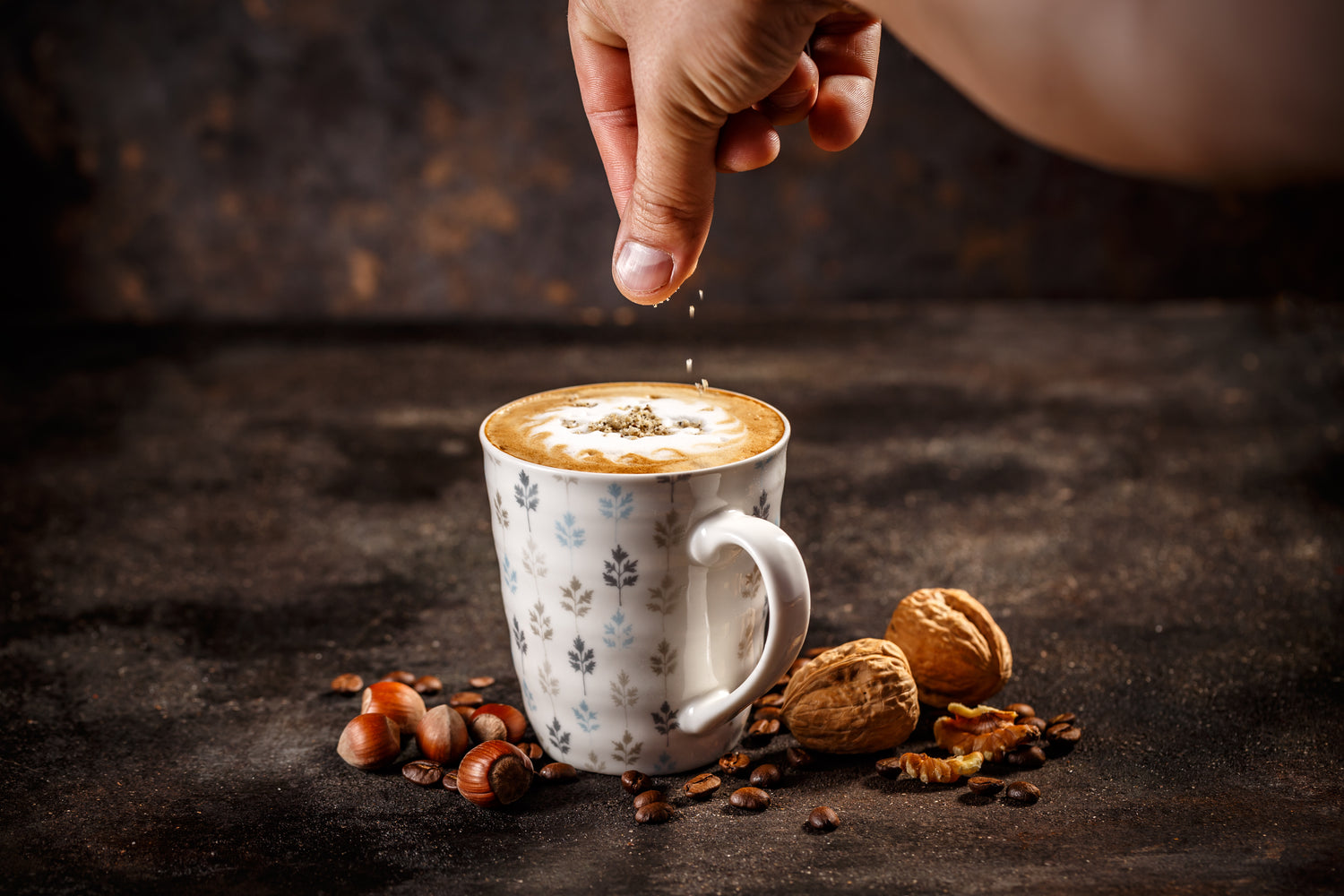 A hand sprinkles toppings onto a frothy coffee in a patterned mug, surrounded by coffee beans, hazelnuts, and walnuts on a dark surface.
