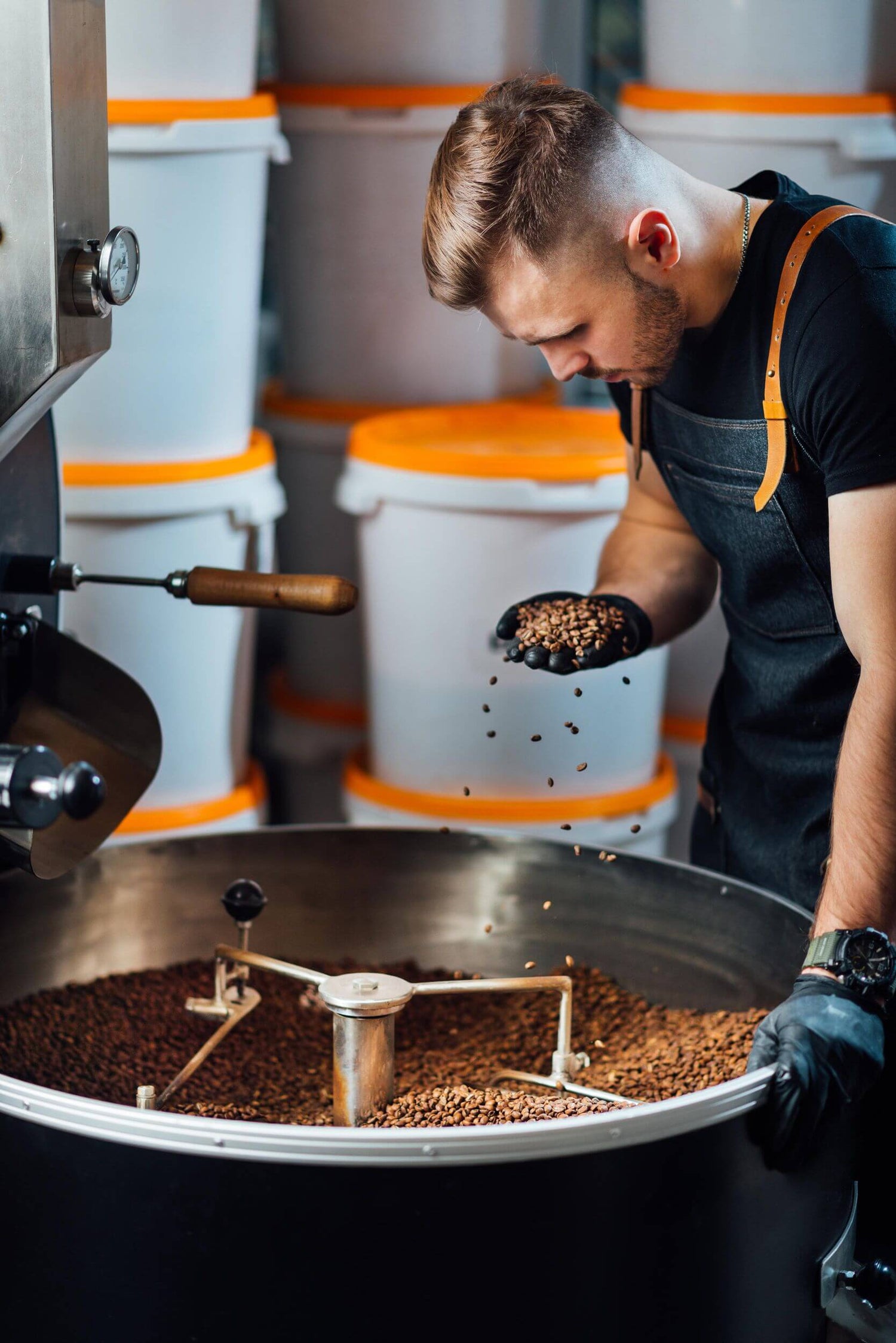 A person wearing black gloves and an apron examines roasted coffee beans by hand over a large industrial roasting machine.